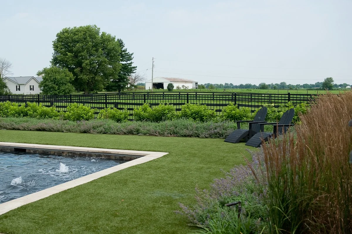 A backyard with a swimming pool, lush green grass, two black Adirondack chairs, a flower bed, a black fence, and a rural landscape with trees, barns, and a field in the distance.