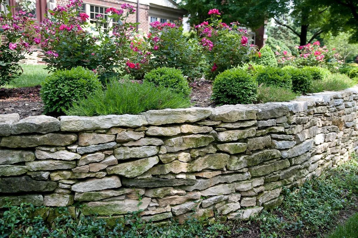 A stone retaining wall with lush green bushes and flowering plants behind it, in front of a residential house surrounded by trees.