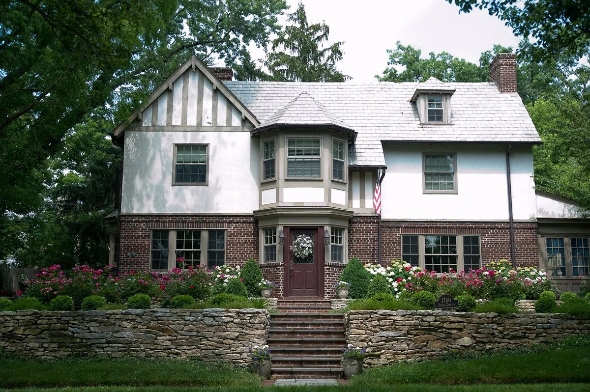 A large two-story house with a brick base and white upper walls, surrounded by a garden with colorful flowers and shrubs, stone steps leading to the front door, and tall trees in the background.