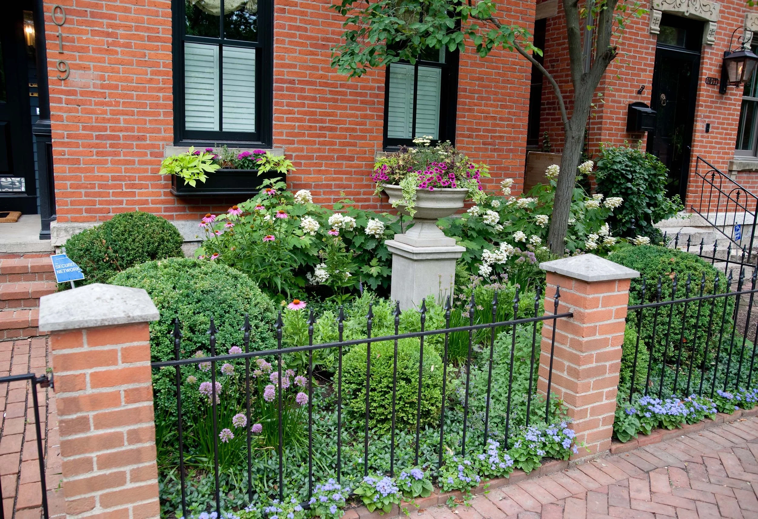 A brick house with two windows and a black door, decorated with flower boxes and surrounded by a small iron fence. The garden includes a variety of colorful flowers and a small tree.