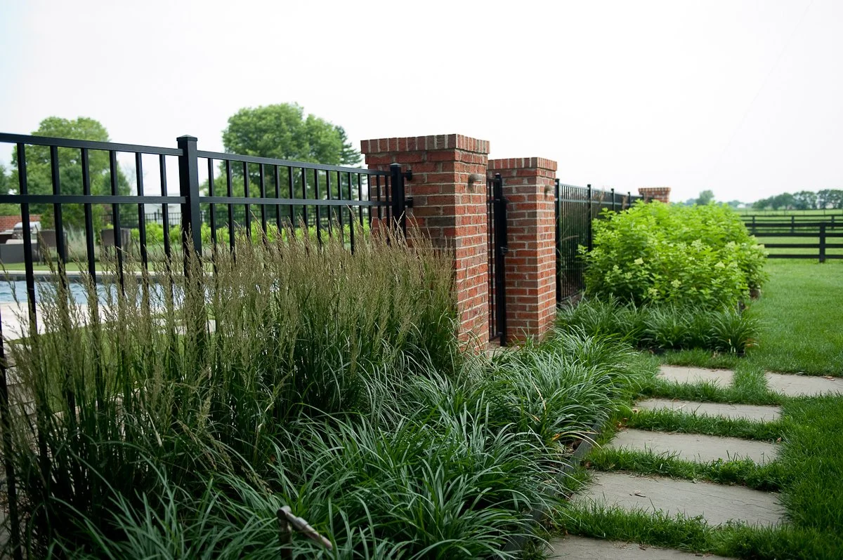A landscaped backyard with a black metal fence, brick pillars, a grassy lawn, stone pathway, and lush green plants.