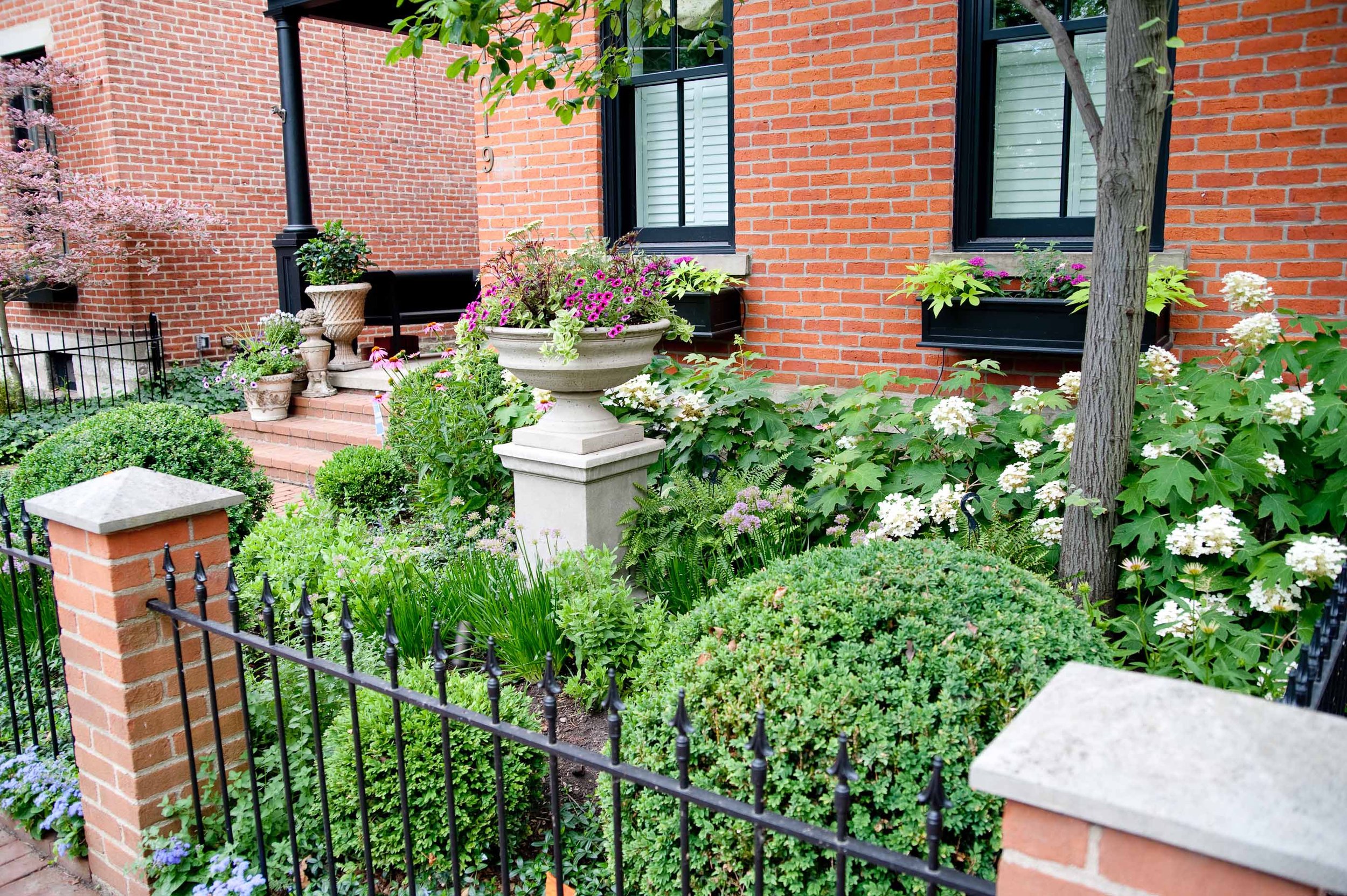 A garden with flowering plants and shrubs in front of a red brick house with black-framed windows. There is a black fence and a brick pathway with steps leading to the house entrance.