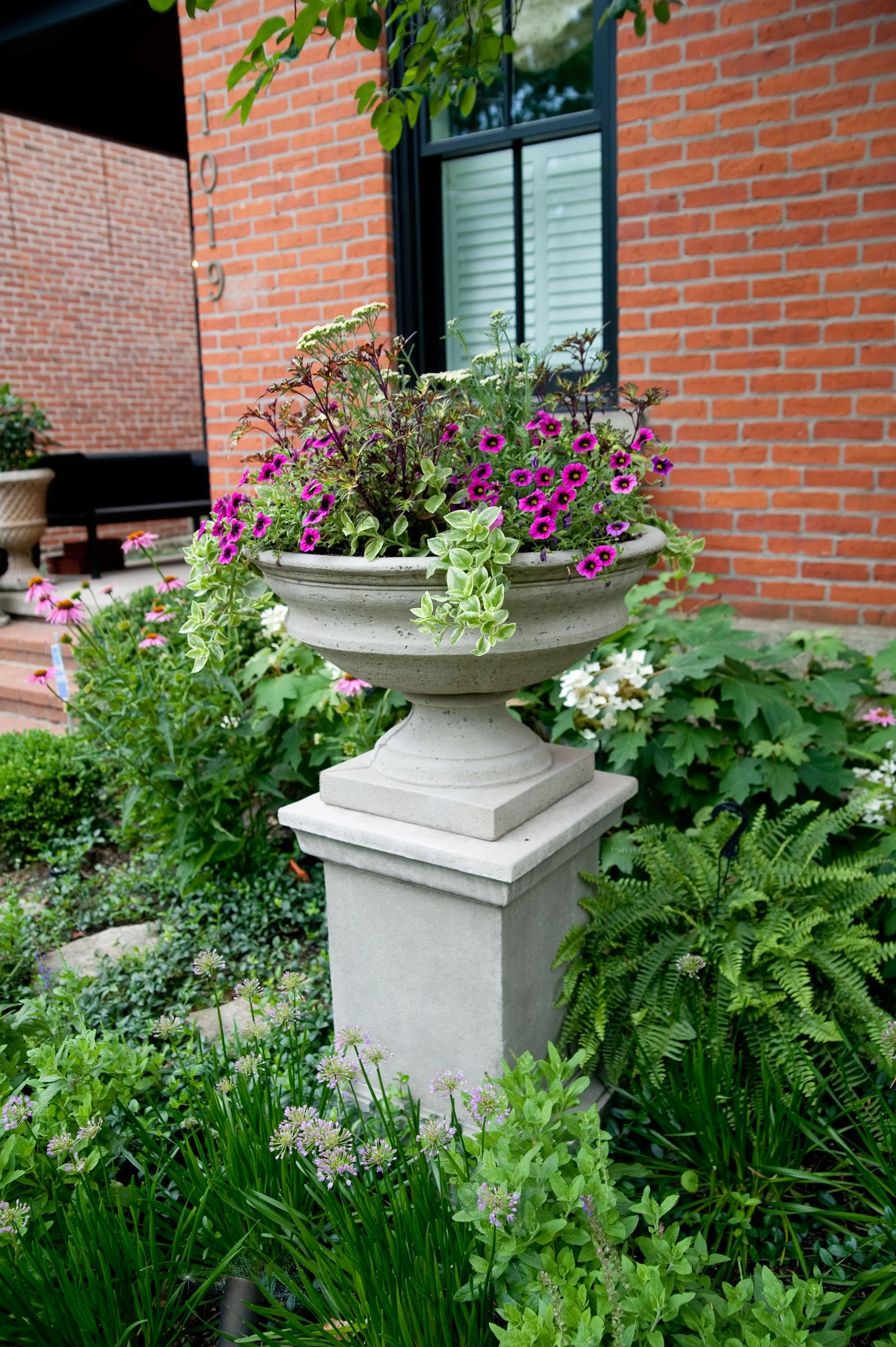 A garden with various green plants and purple flowers in a decorative stone planter on a tall pedestal in front of a brick house with a window.