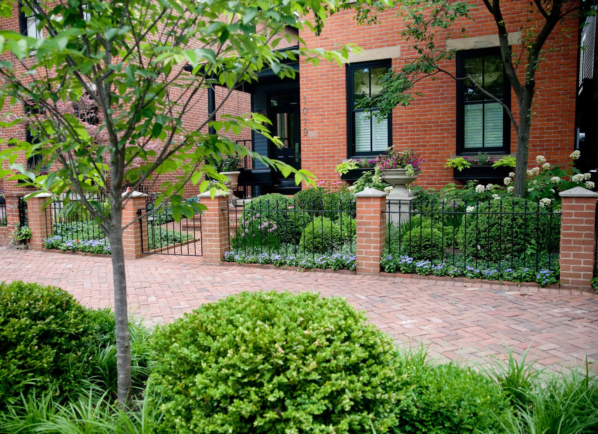 Front yard garden with brick pathway, lush green bushes, trees, and flowering plants in pots near a brick house with black-framed windows.