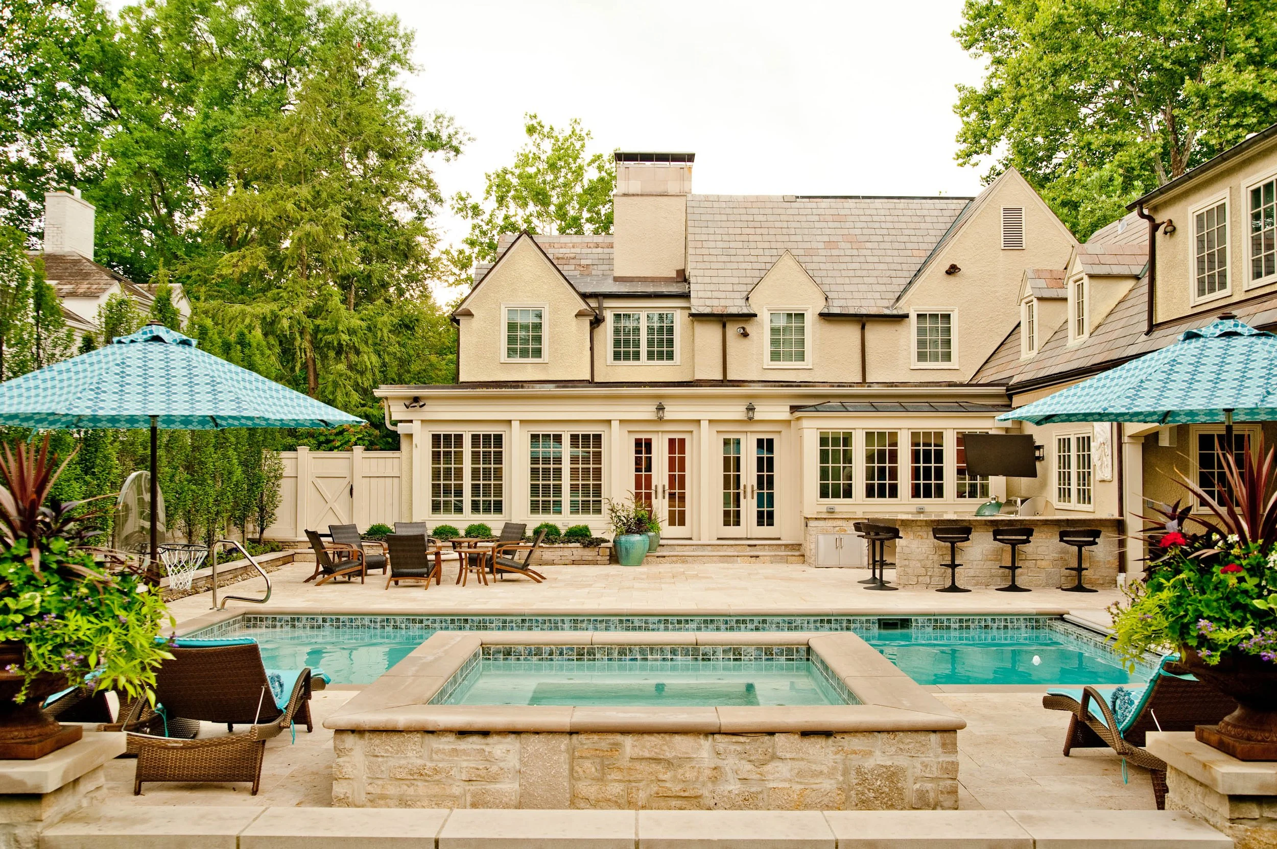 Backyard patio with swimming pool, outdoor furniture, umbrellas, food prep area, and a large house with multiple windows and steeply pitched roofs, surrounded by green trees.