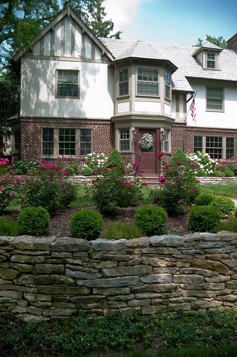 A large two-story house with a brick and white exterior, surrounded by a lush garden and a stone wall in the foreground.