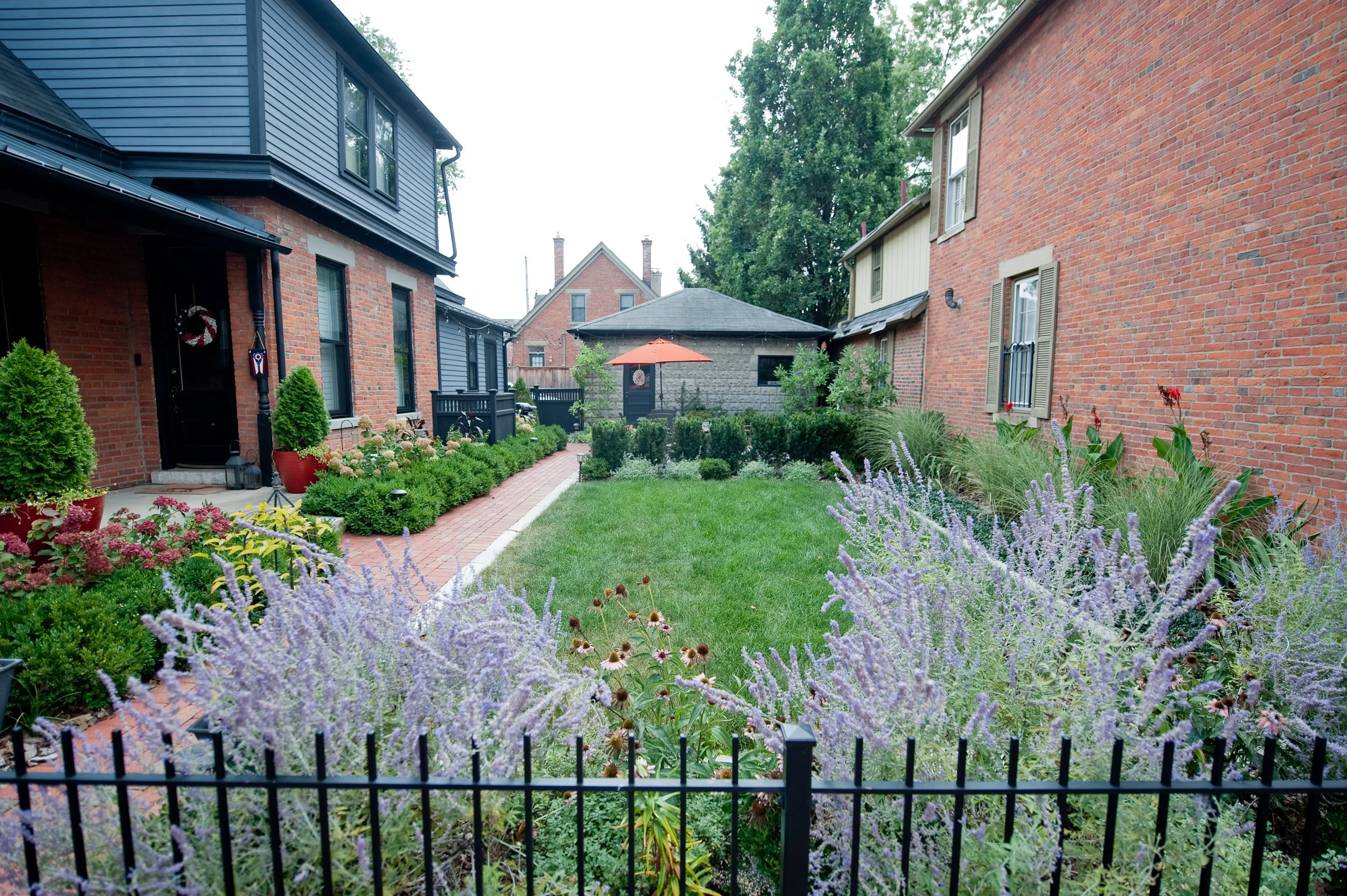 A backyard garden with a brick house on the right and a blue house on the left, featuring a brick pathway, lush green plants, colorful flowers, and an orange patio umbrella.