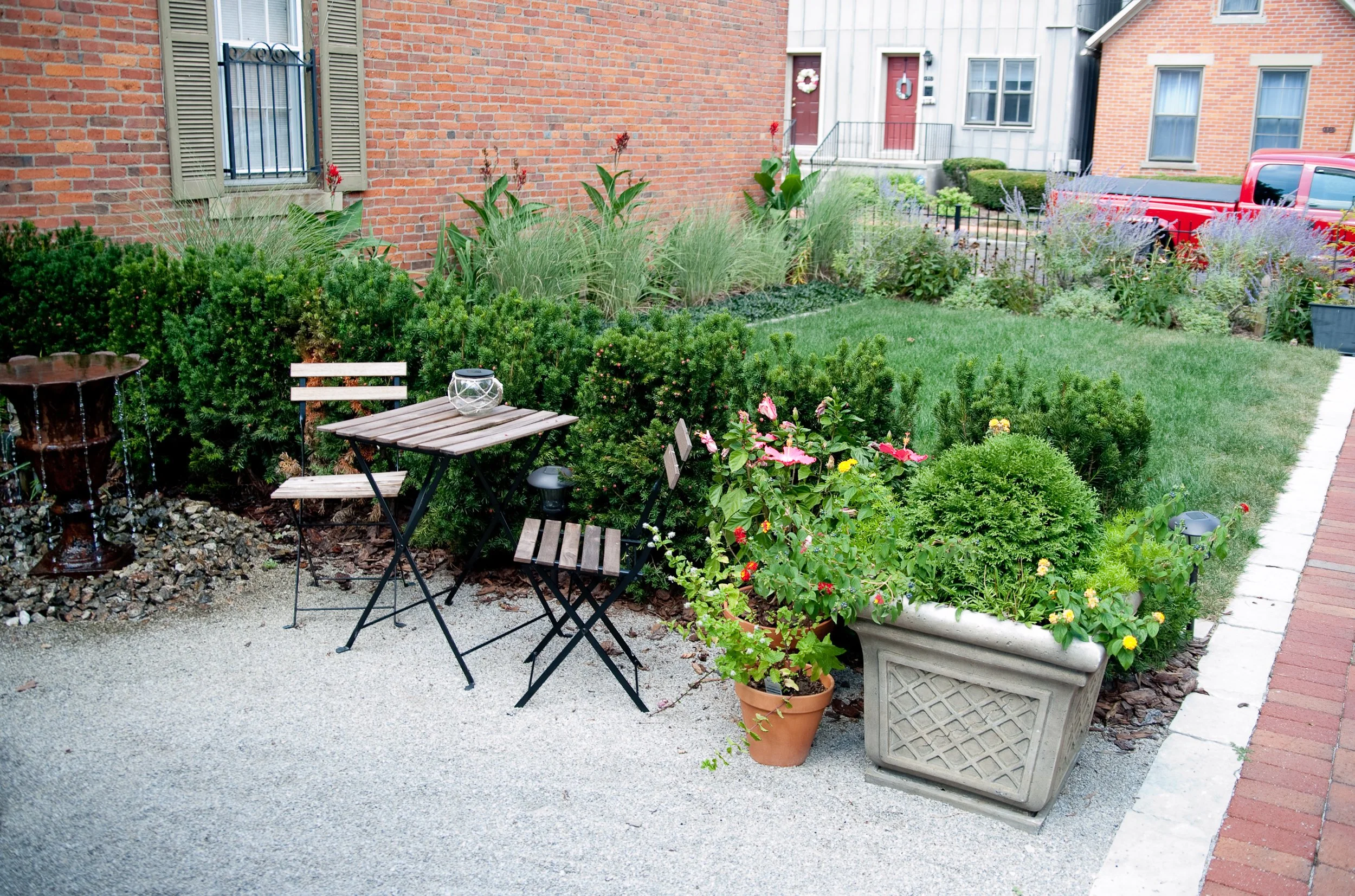 A small outdoor garden with various plants and flowers, a gravel seating area with a wooden table and chairs, and a large decorative planter, adjacent to a brick pathway and residential buildings.