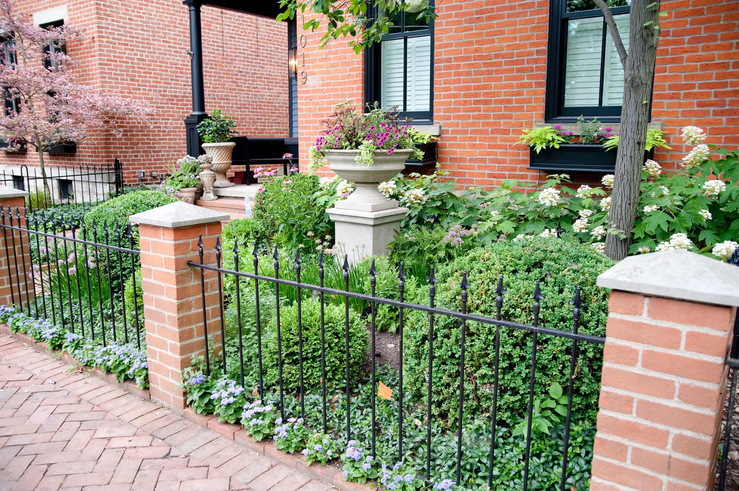 A brick house with black window frames, decorated with colorful flowers and green plants in planters, surrounded by a black metal fence and a brick sidewalk.