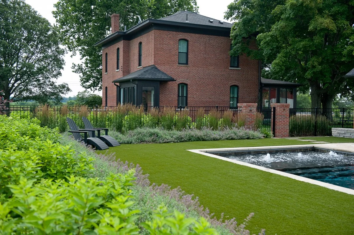 A backyard with a swimming pool, two black lounge chairs, lush greenery, and a brick house surrounded by a black metal fence.