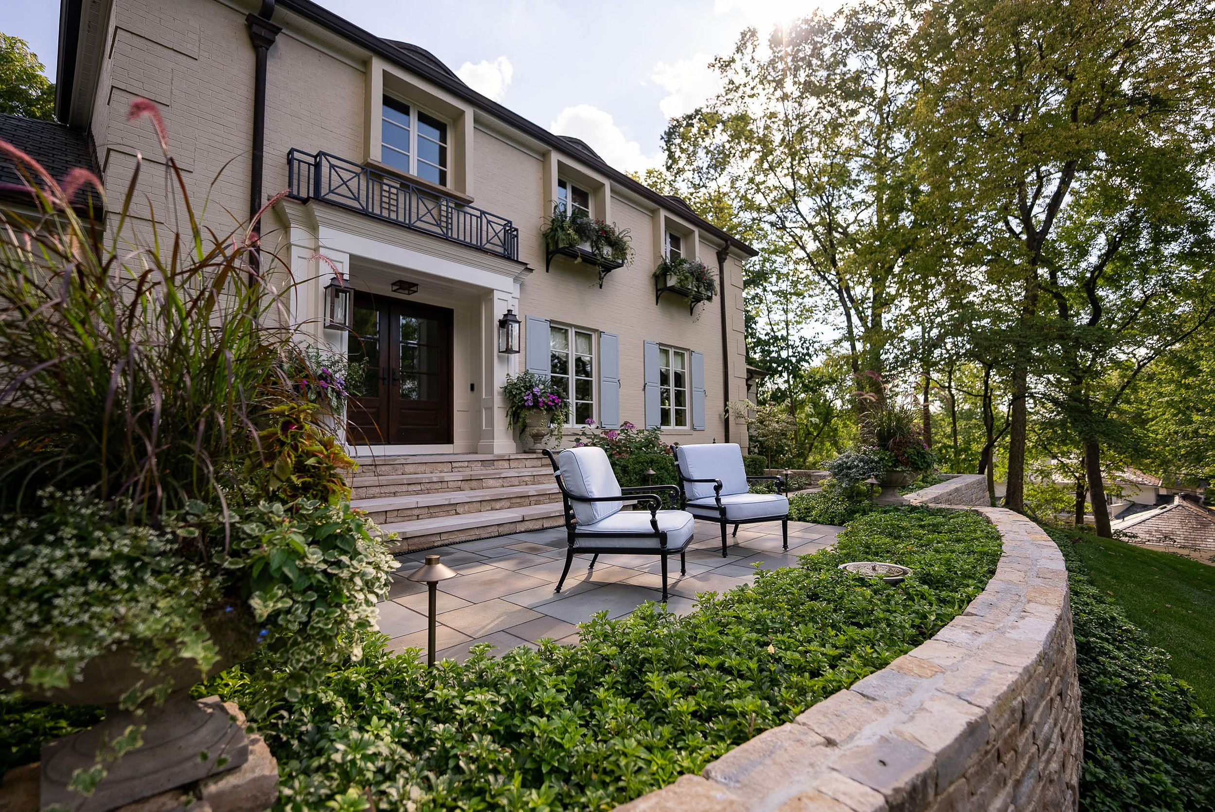 A house with a brick exterior and black metal balcony railings, surrounded by trees and a landscaped yard with outdoor seating and potted plants.