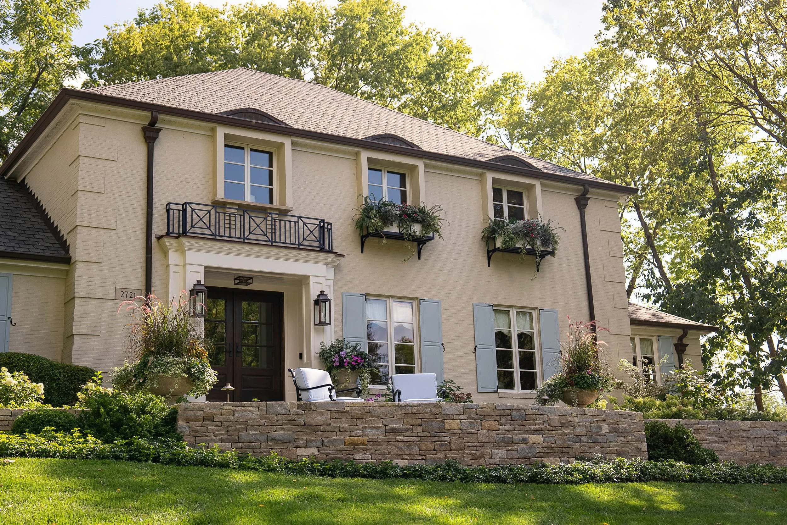 Front view of a two-story cream-colored house with blue shutters, flower boxes, a balcony, and a landscaped front yard with patio furniture, trees, and a stone retaining wall.