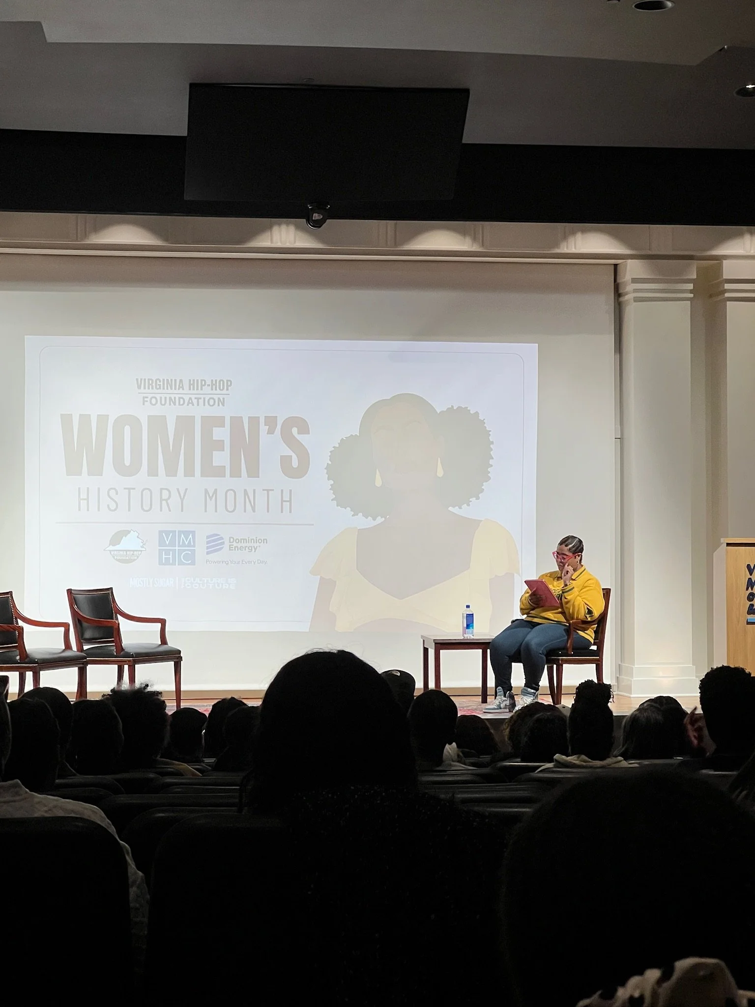 A woman in a yellow jacket and glasses sitting on a stage chair, reading a book, during a Women's History Month event organized by the Virginia Hip-Hop Foundation. Audience members are visible in the foreground.