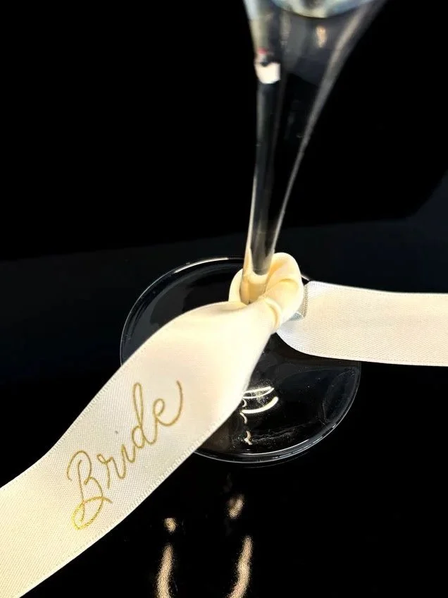 Guldfolierad text "bride" på vitt sidenband som är knytet runt ett champagneglas