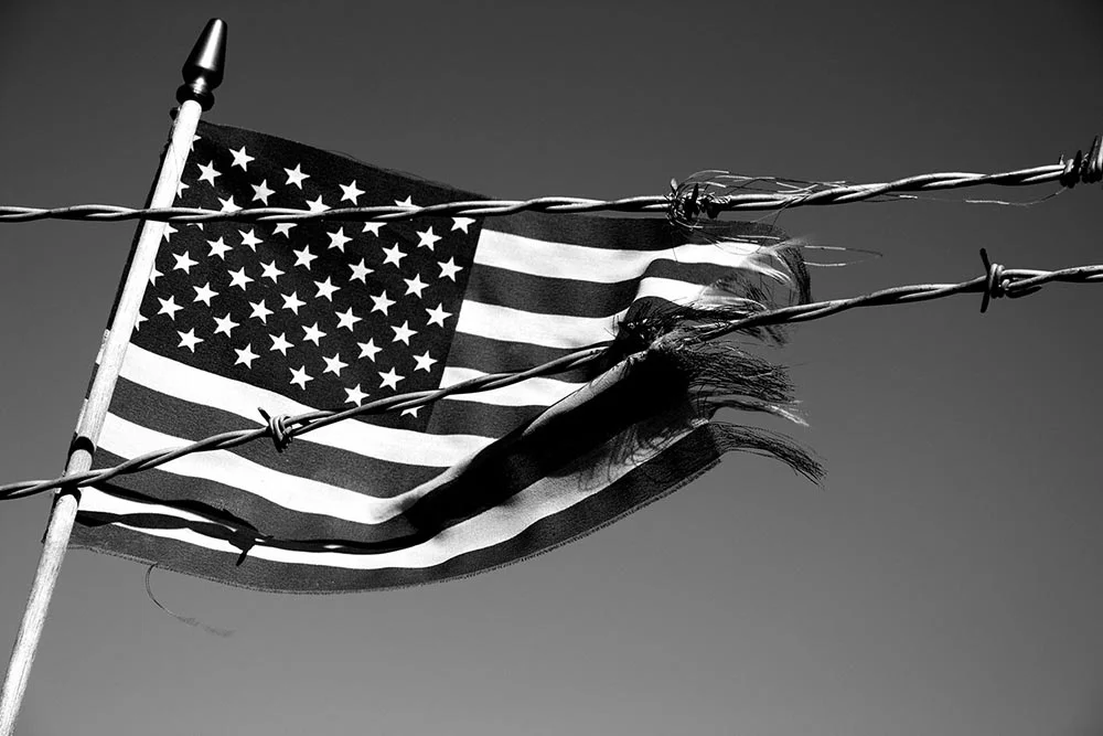 Black and white photo of an American flag being ripped apart by barbed wire.