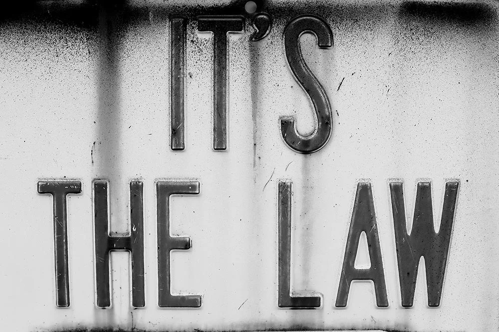 Black and white photo of a rusted sign that says "It's the Law."