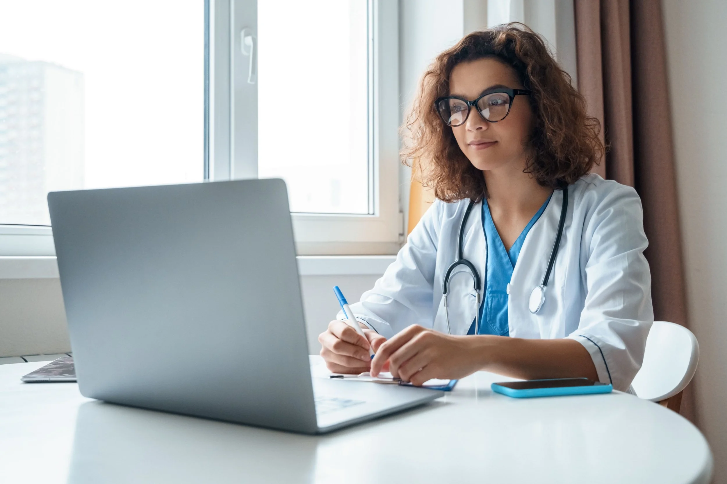 A female doctor with curly hair and glasses sitting at a desk, working on a laptop, with a stethoscope around her neck, in a bright room near a window.