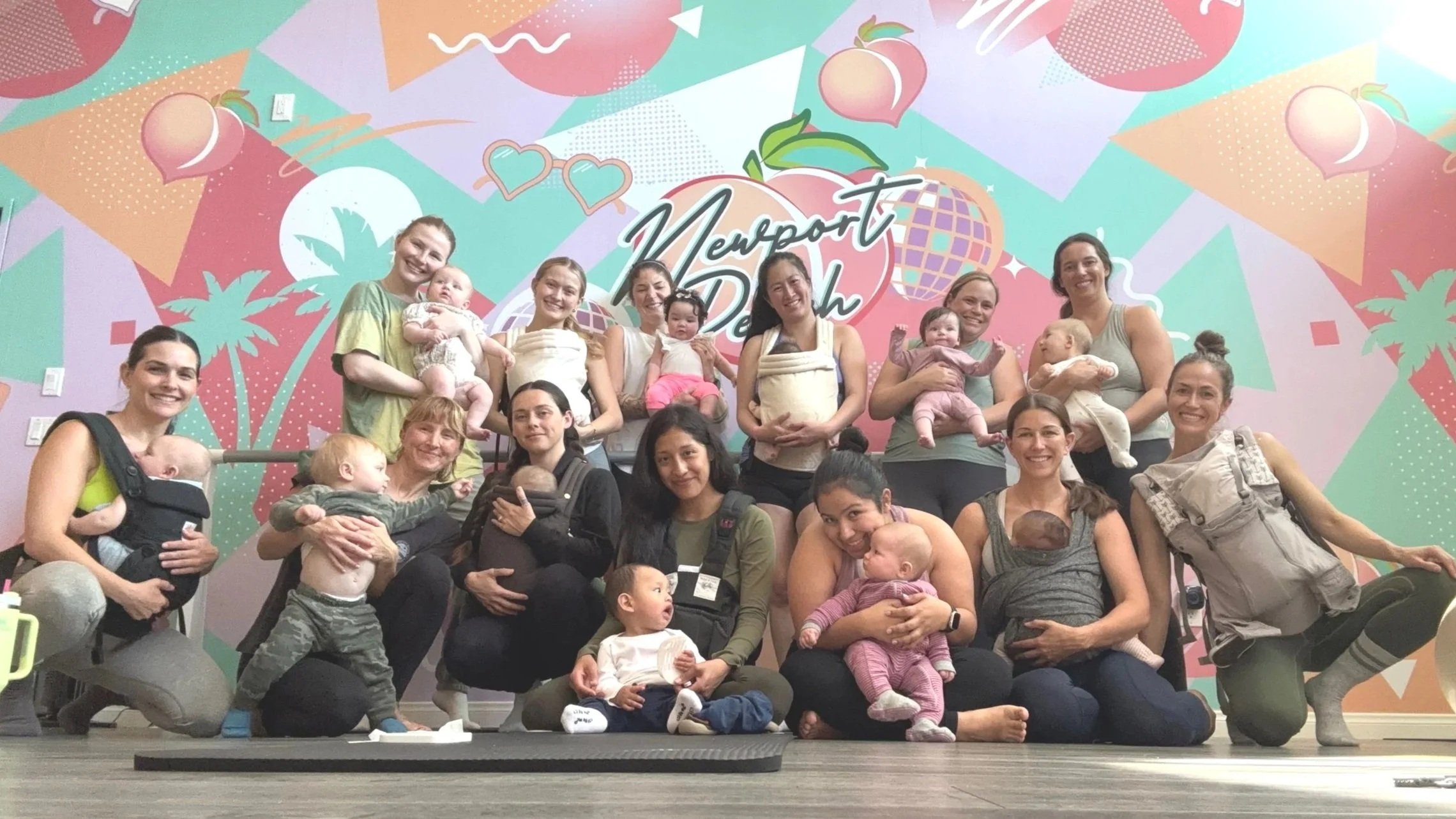 Group of women and children, some in carriers, posing in front of a colorful mural with the text 'Newport Baby Detox' and playful graphics of peaches, hearts, and geometric shapes.