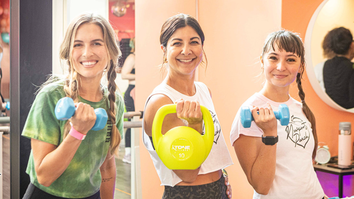Three women smiling at a gym, holding dumbbells and a kettlebell during a workout.
