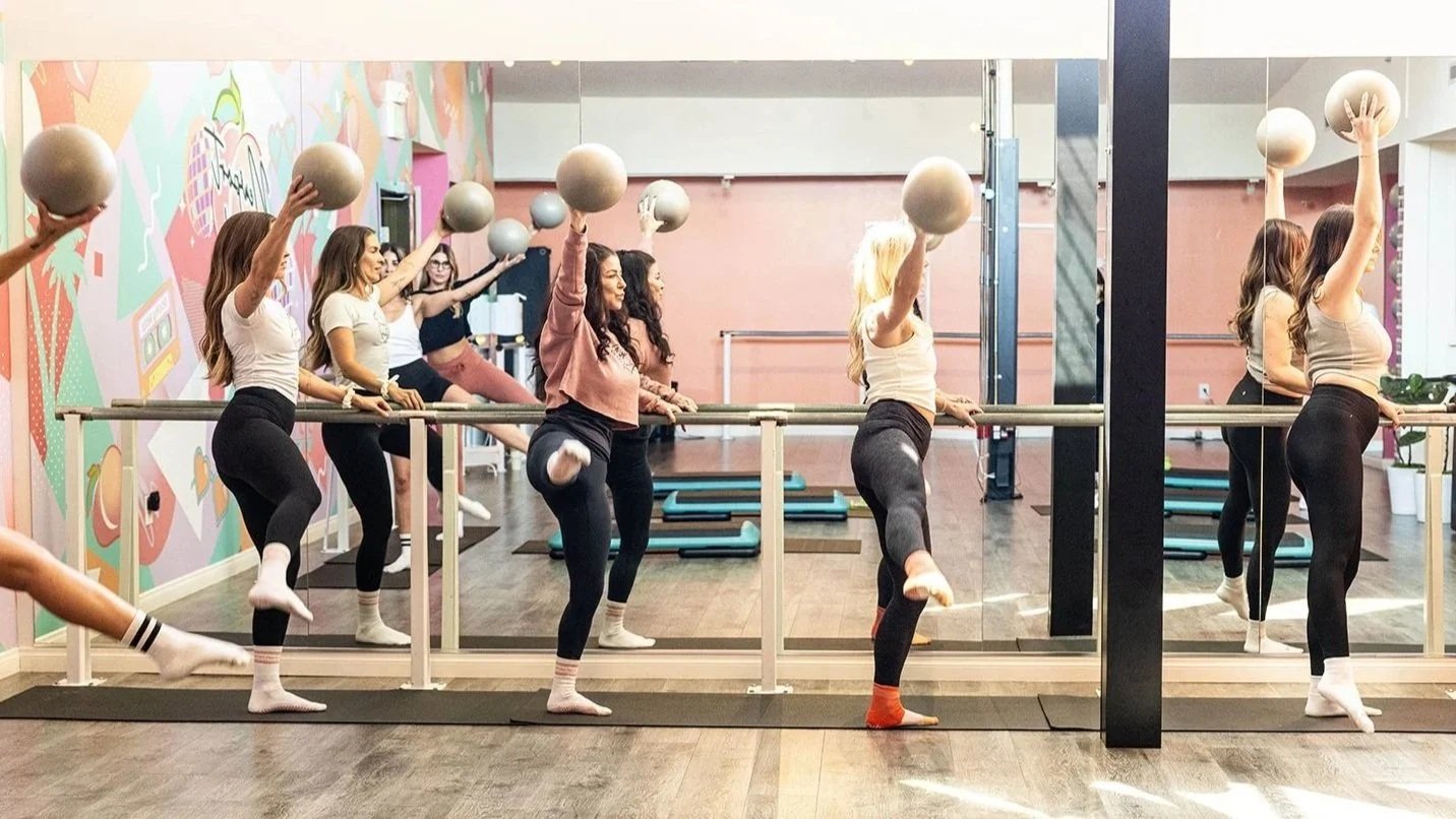 A group of women attending a barre class in a fitness studio, lifting grey exercise balls overhead while balancing on one leg with the other leg bent.