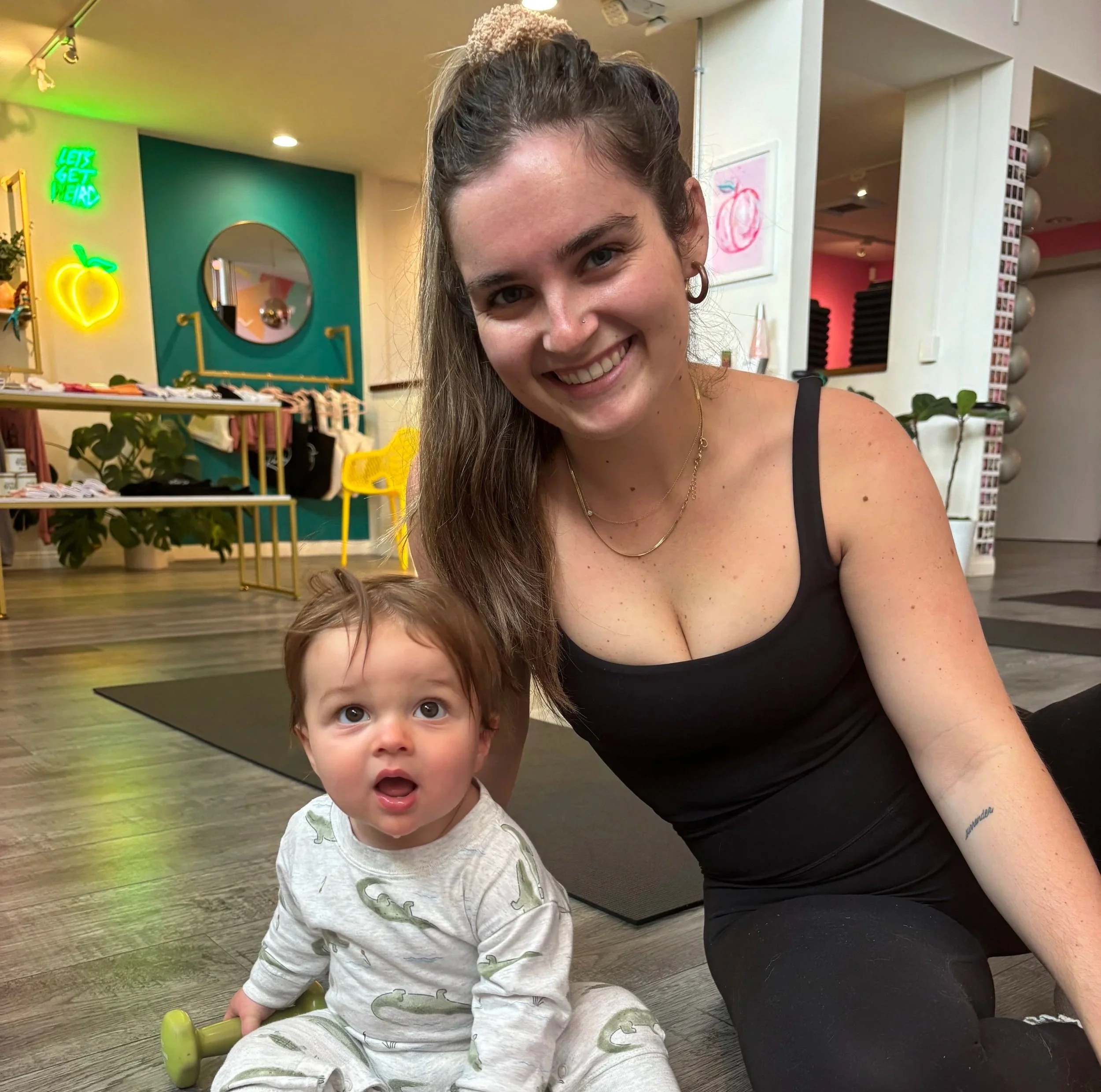 A young woman with long brown hair, wearing a black top and gold jewelry, smiling at the camera, seated next to a young boy sitting on the floor, wearing pajamas with a dinosaur print, in a colorful indoor setting with neon signs and decor.