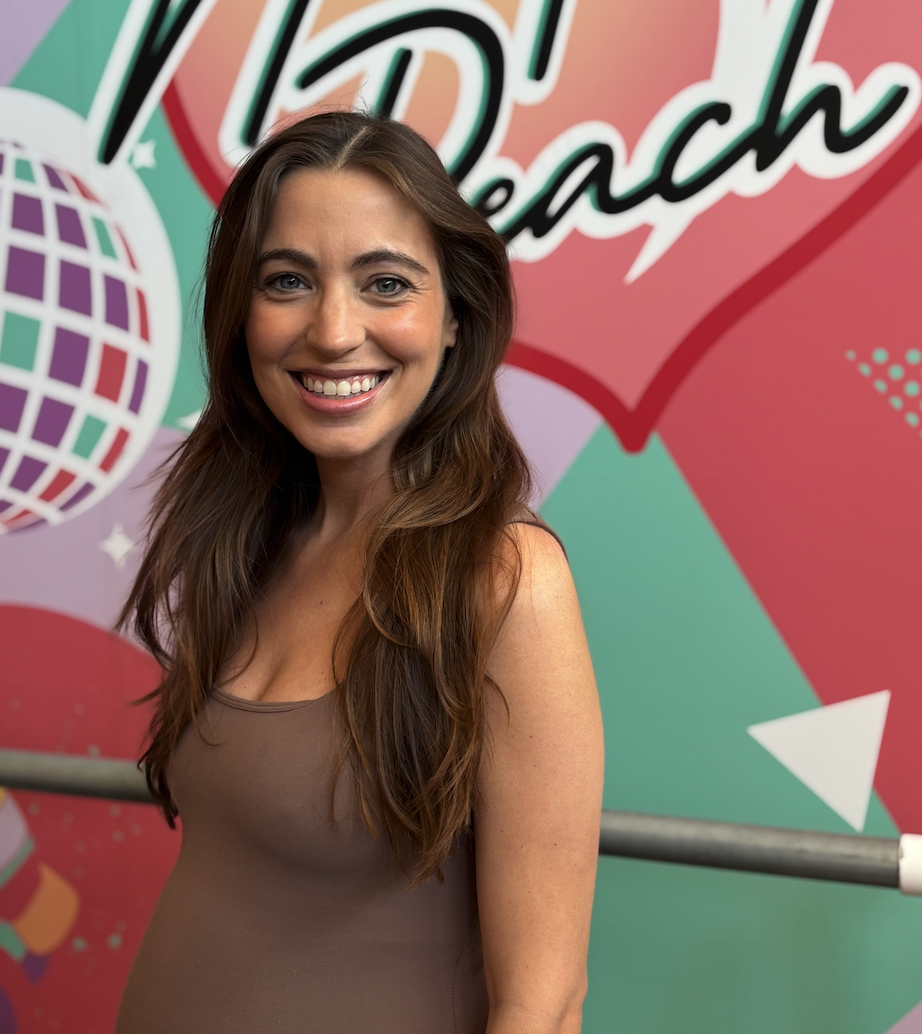 A woman with long brown hair smiling in front of a colorful backdrop with the words 'BI Beaches' and a disco ball design.