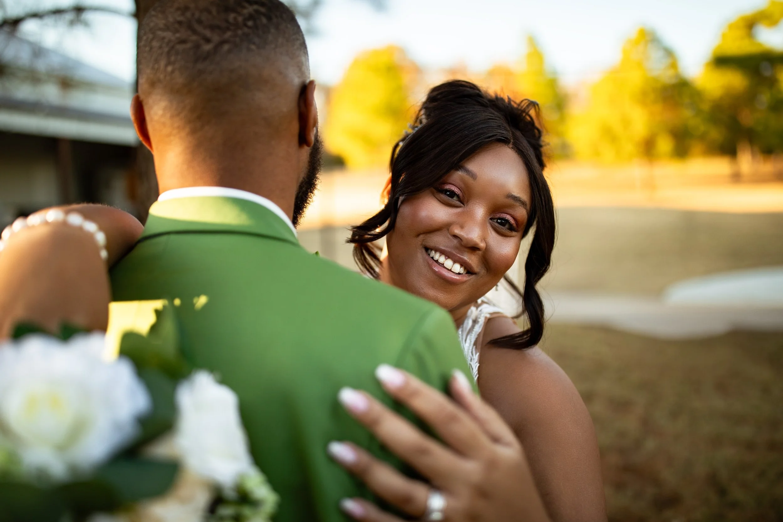 A woman smiling and hugging a man dressed in a green suit outdoors during sunset, with trees and a grassy area in the background.