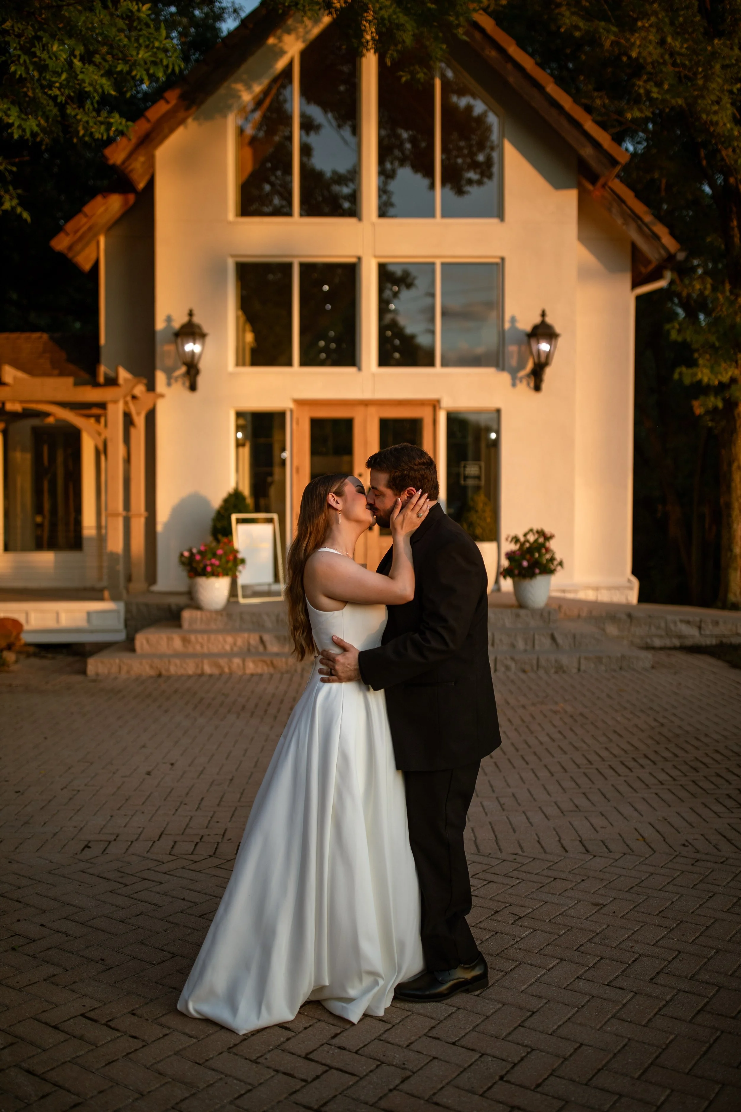 A bride and groom kissing in front of a large house at sunset.