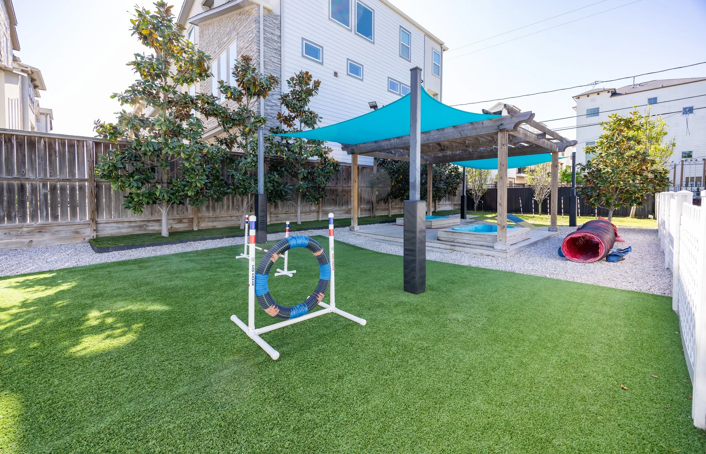 Backyard with artificial grass, agility ring, dog tunnel, small pool, shade canopy, trees, and a wooden fence.