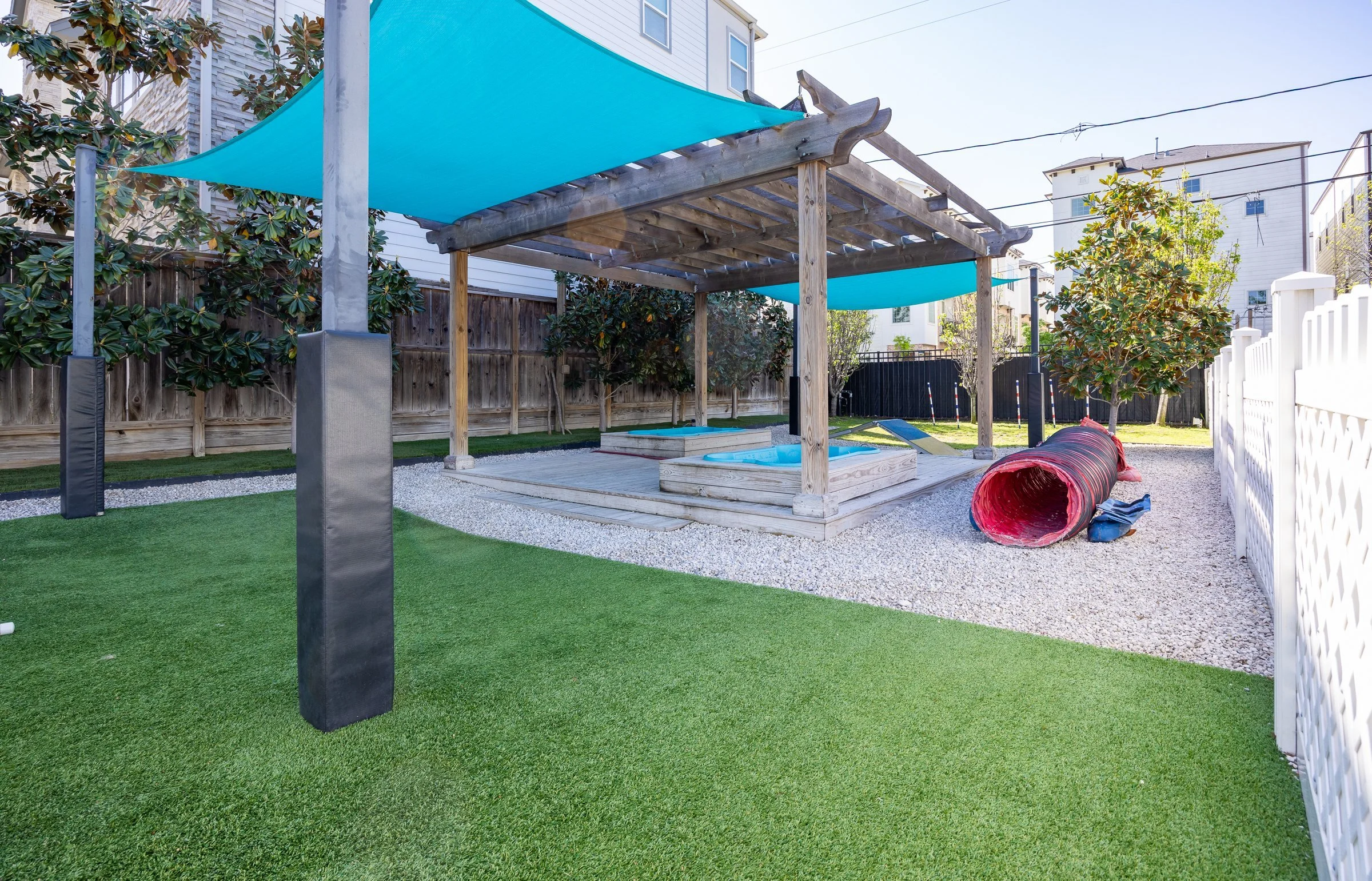 Backyard with artificial grass, wooden shade structure, small hot tub, green trees, slide, tunnel, and white fence.