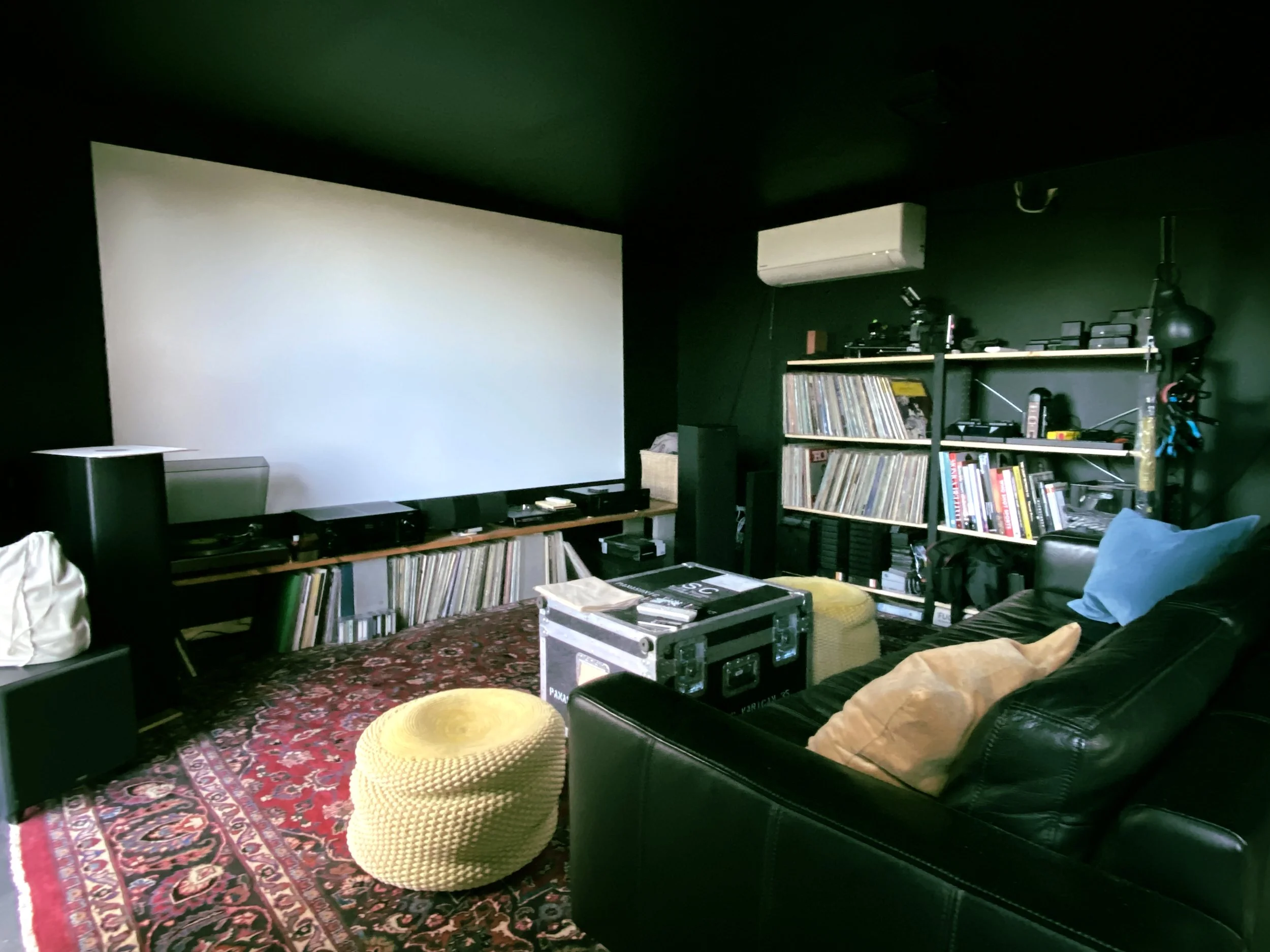 A room with a large white screen, black walls, a black leather couch with a throw pillow, a yellow pouf, a bookshelf filled with vinyl records and various electronic equipment, an air conditioning unit, and audio/video equipment on a low black stand with a patterned rug on the floor.