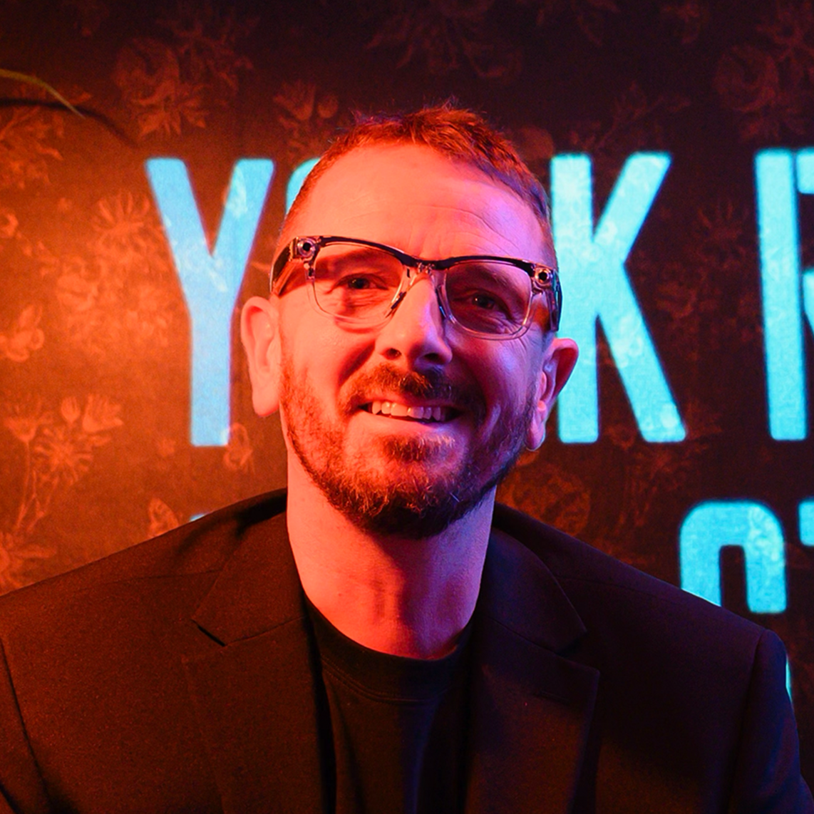A man with glasses, a beard, and a mustache, dressed in a dark suit, is smiling. The background is dark with blue vertical lines of light.