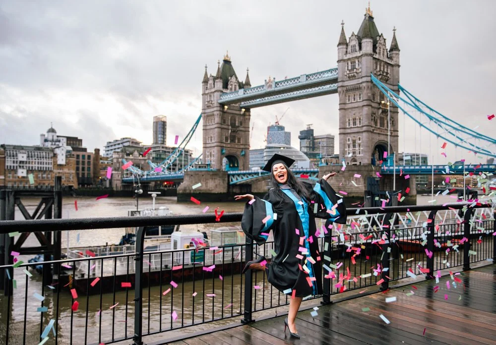 A woman in graduation gown and cap celebrating on a riverside promenade with Tower Bridge in London in the background, surrounded by pink and blue confetti. Captured by a London photographer