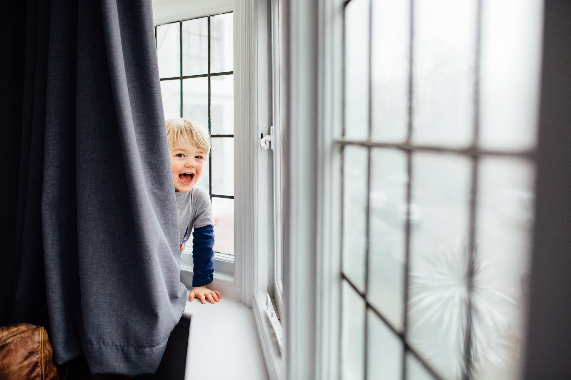 A young boy with blond hair smiling and leaning out from behind a curtain by a window in his home. Captured by a London Family photographer