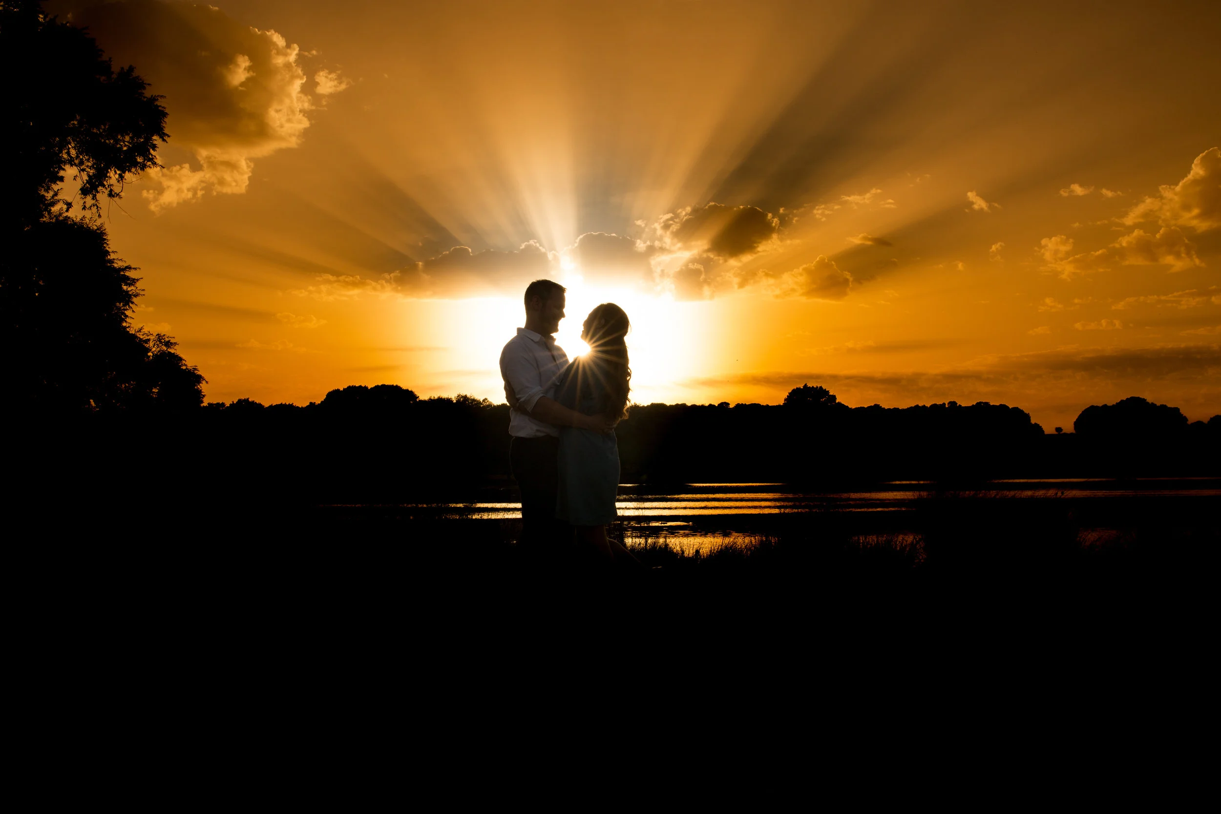 A silhouette of a couple embracing at sunset near a body of water, with the sky painted in orange and yellow hues and clouds scattered across the sky. Captured by a London Engagement photographer
