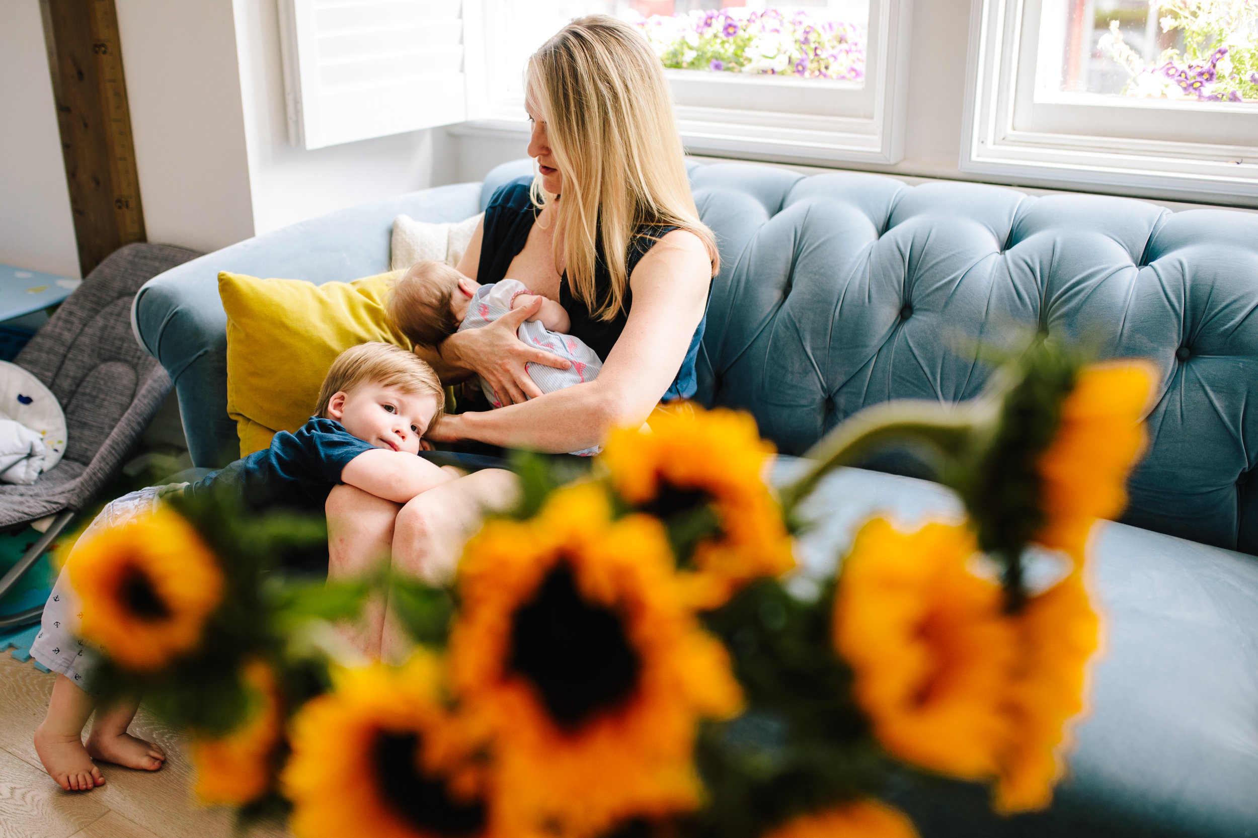 A woman with blonde hair sitting on a light blue couch, breastfeeding a baby, with a young boy leaning against her leg. The foreground has yellow and orange flowers, slightly out of focus. Captured by a London Family photographer