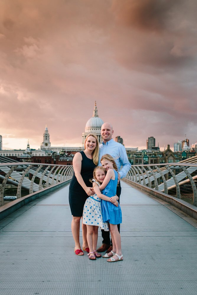 Family of four, two adults and two children, standing on Millennium Bridge in London, with St. Paul's Cathedral in the background during sunset. Captured by a London Family photographer