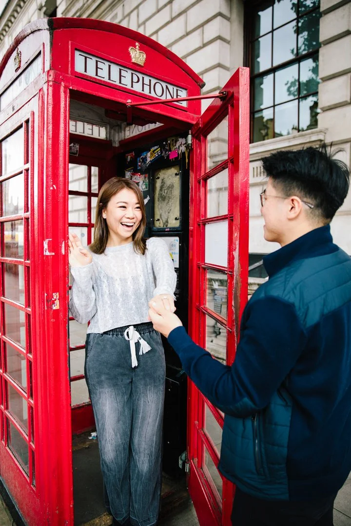 A smiling woman on a red British telephone booth talking to a man outside on a city sidewalk.