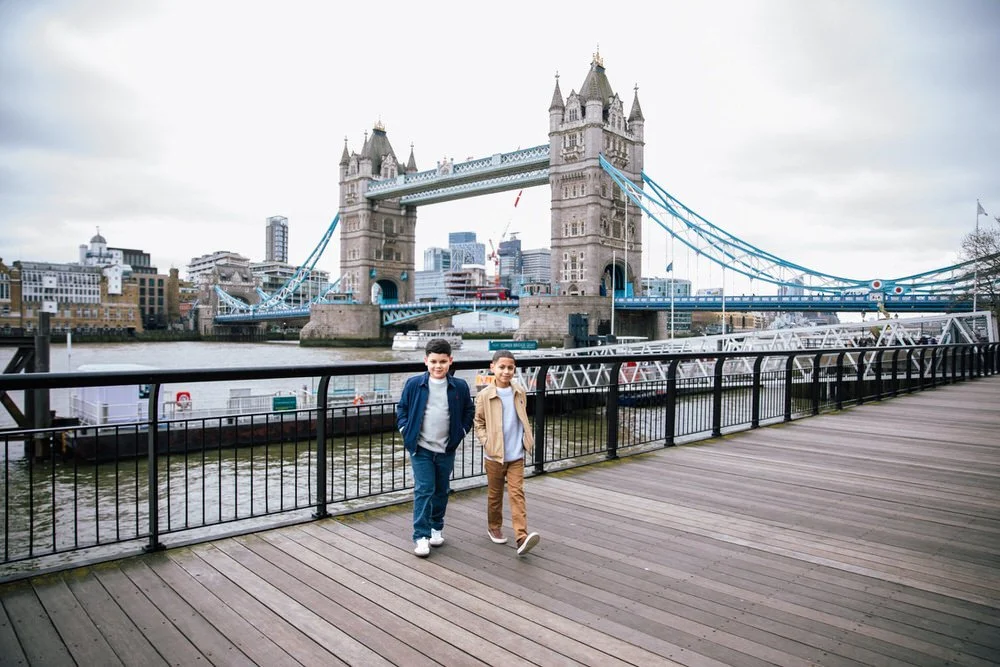 Two boys stand on a wooden boardwalk by the Thames River in London, with Tower Bridge in the background. Captured by a London Family photographer