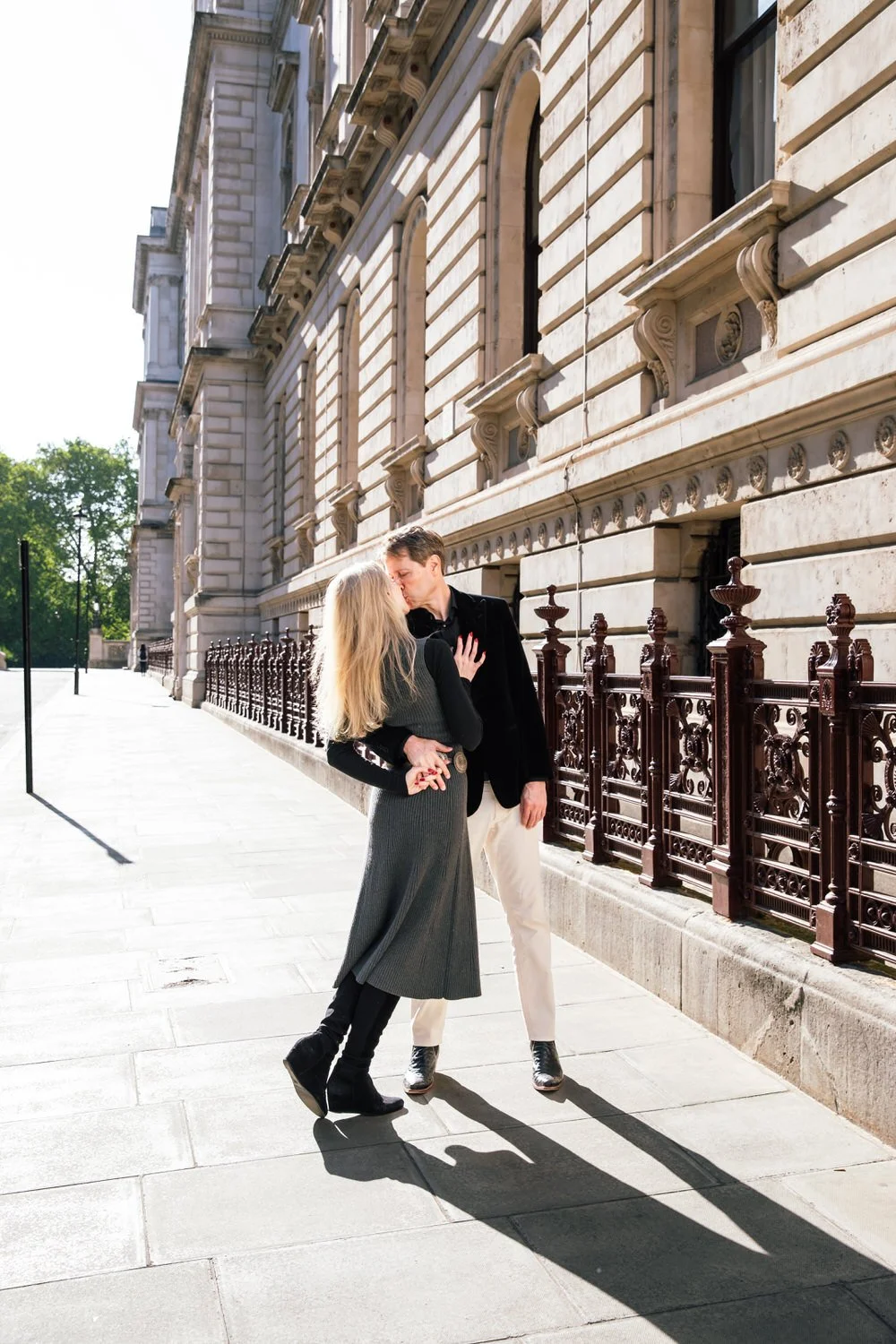 A man and woman kissing on a city sidewalk beside a historic stone building with ornate windows and a decorative iron fence. Captured by a London Engagement photographer
