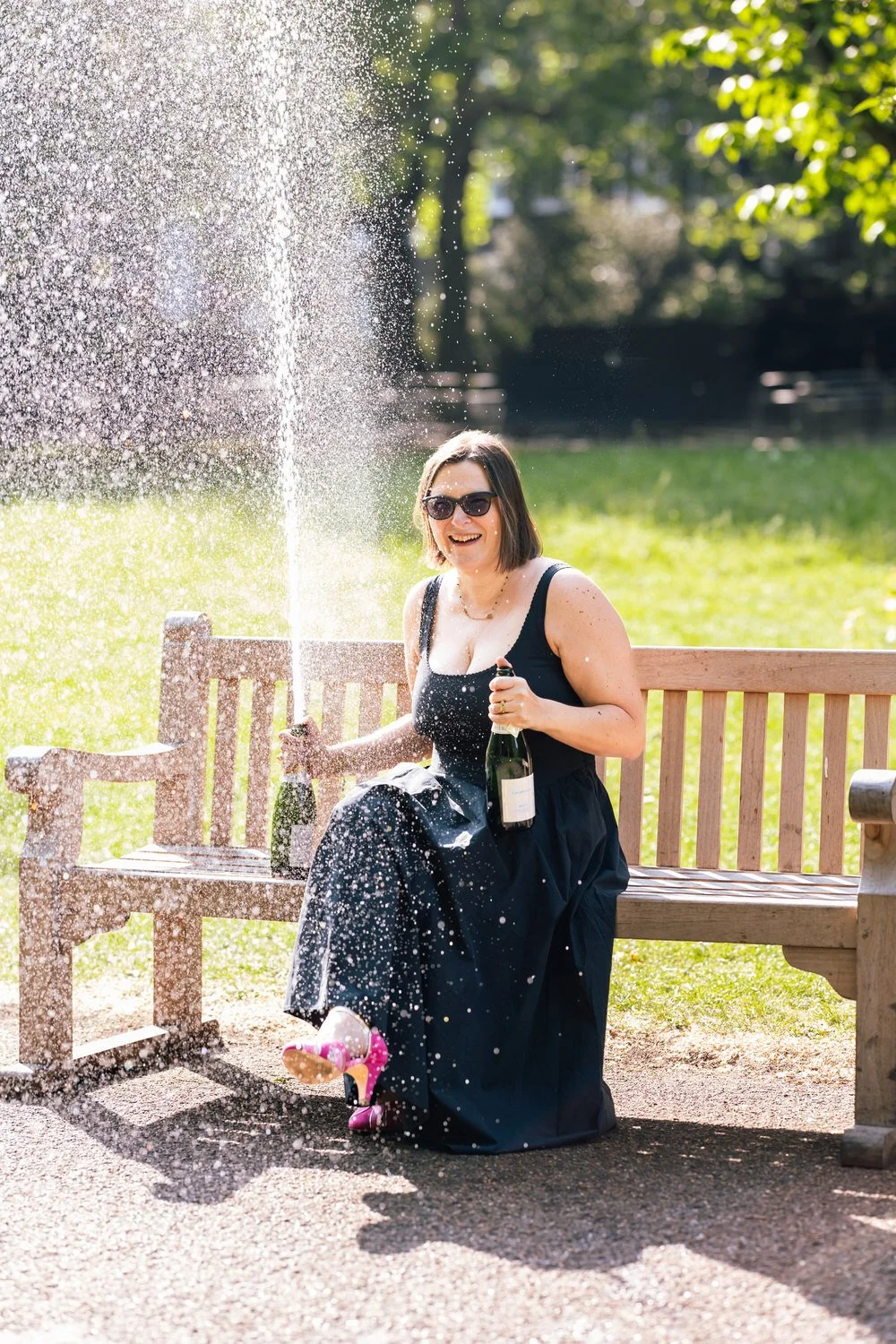 Woman in sunglasses and black dress sitting on a park bench, laughing and spraying champagne, with pink high heels, on a sunny day in a green park. Captured by a London photographer