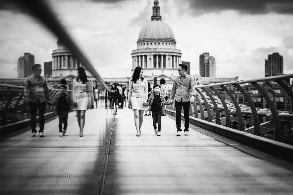 A family of two adults and two children walking on a modern bridge with the St. Paul’s Cathedral in London in the background, black and white photo. Captured by a London Family photographer