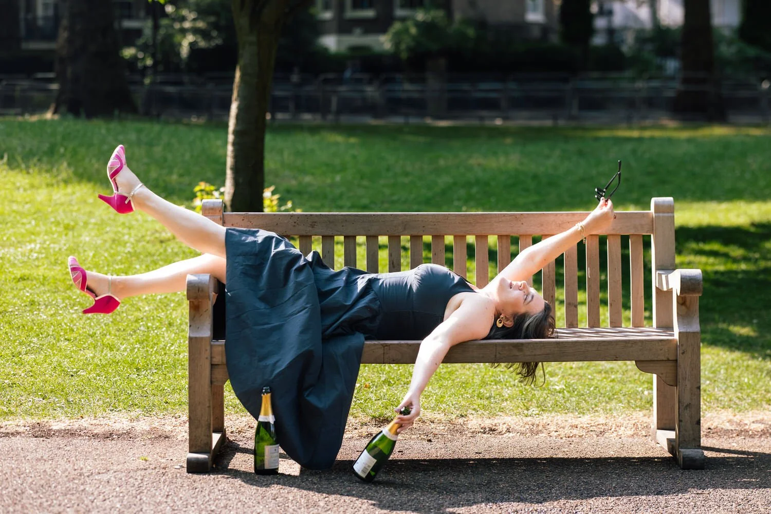A woman in a black dress and pink high heels lies on a park bench, holding sunglasses in one hand and two bottles of champagne placed on the ground beside the bench, in a park with green grass and trees. Captured by a London photographer