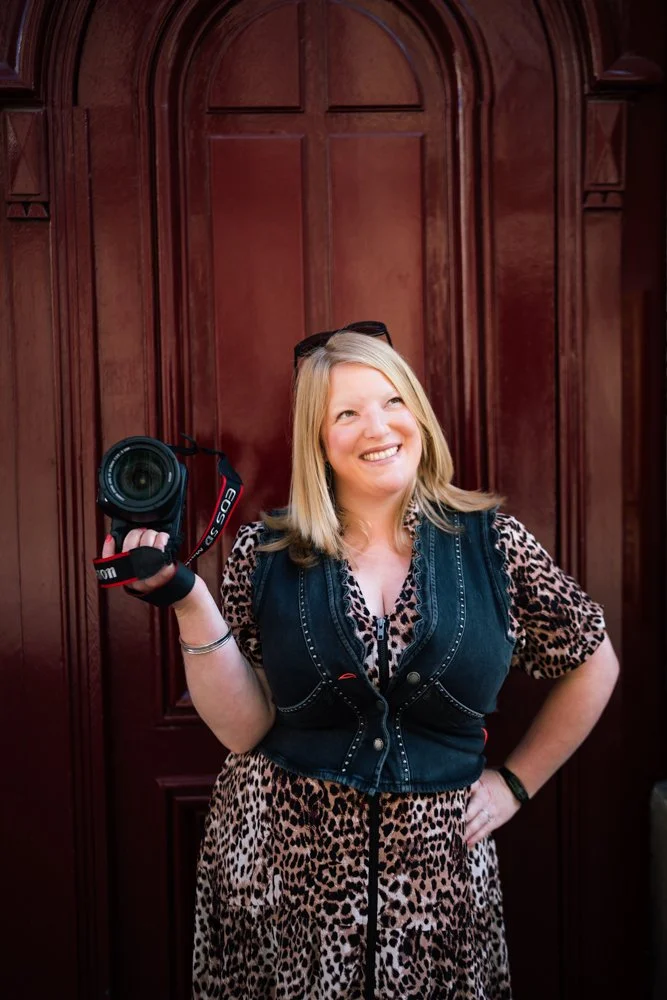 Kirsty Hamilton Photographer holds camera in hand and looks to the upper right of the frame while smiling