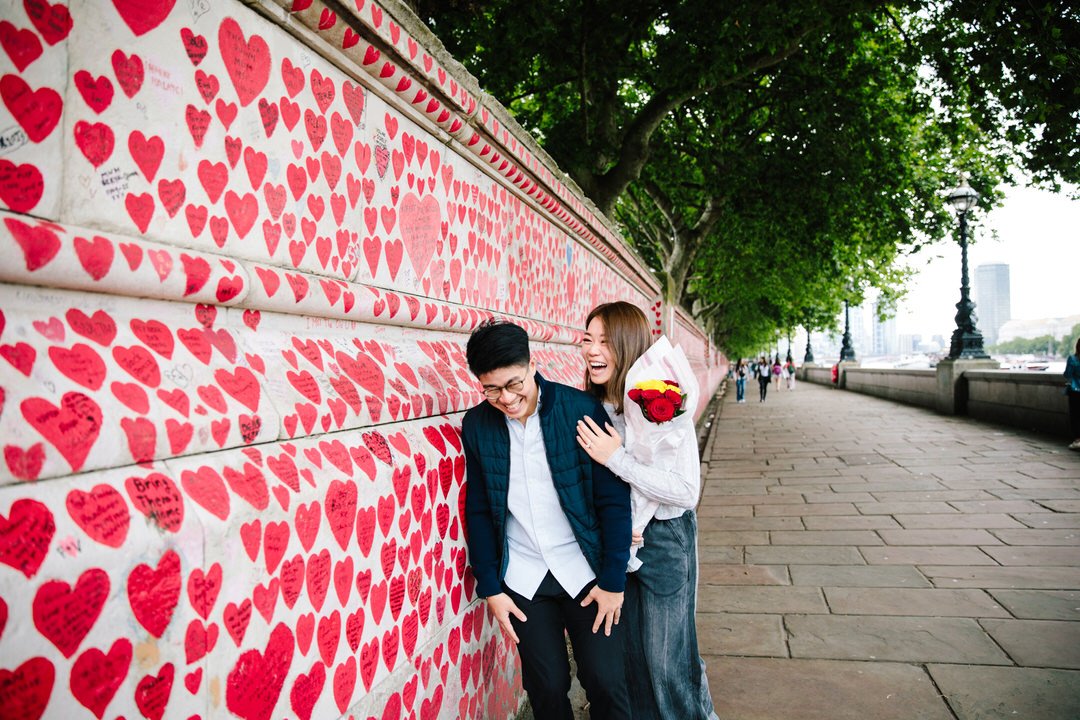 A man and woman are standing near a wall decorated with numerous red paper hearts. The woman is holding a bouquet of red and yellow roses and is smiling, while the man is laughing. Captured by a London Proposal photographer