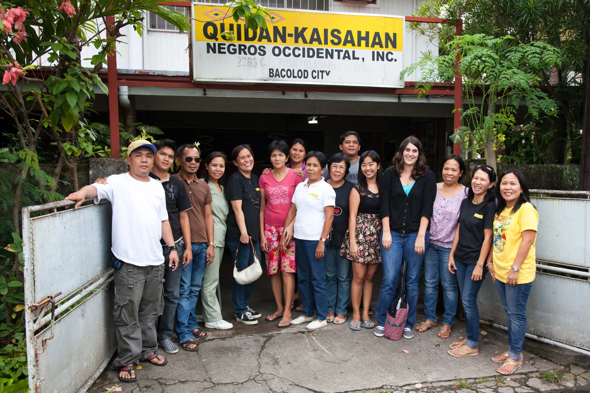Group of people standing outside in front of a sign that reads 'QUIDAD-KAISAHAN NEGROS OCCIDENTAL, INC. BACOLOD CITY'.