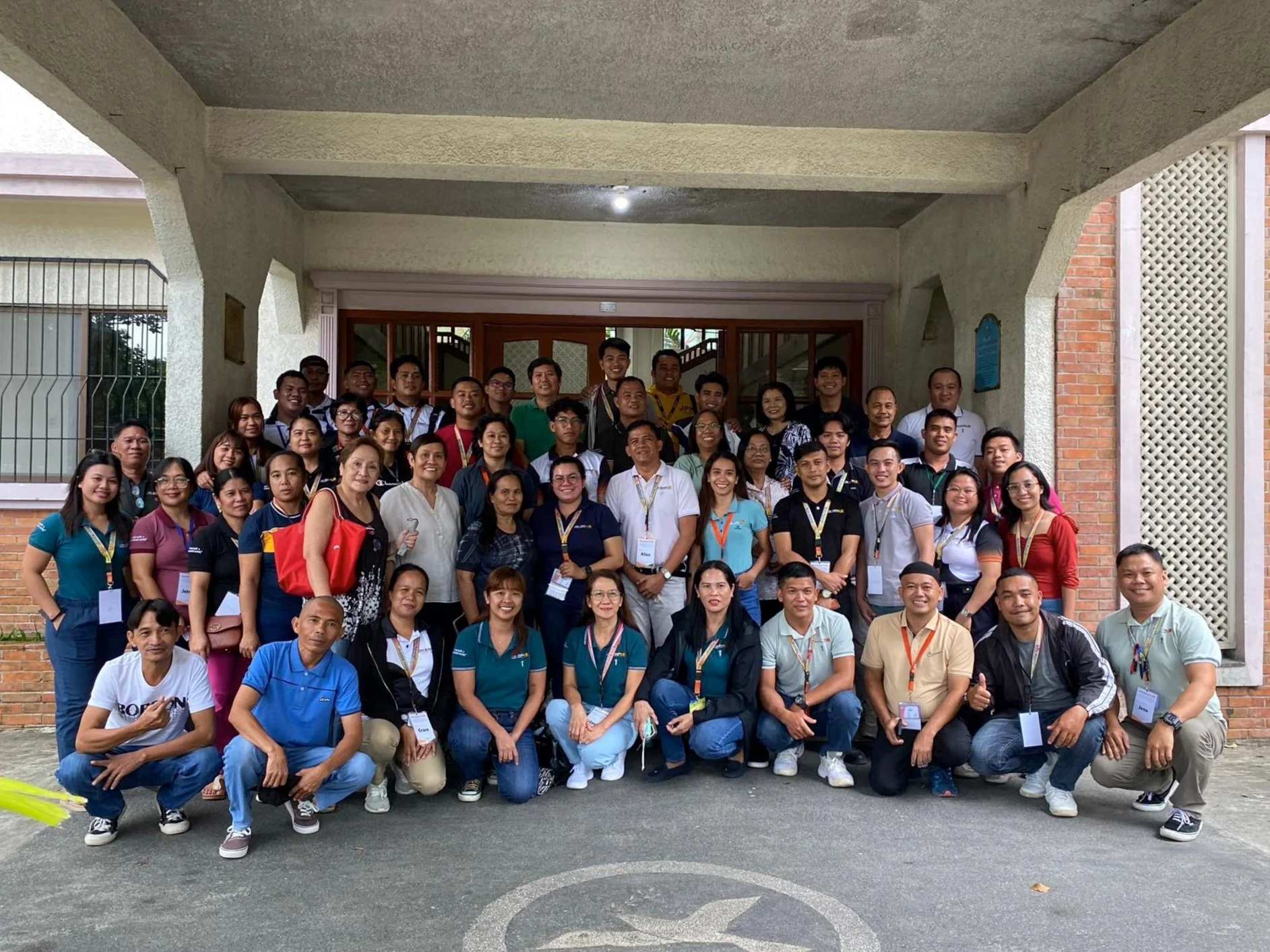 A large group of diverse people posing for a photo outdoors in front of a building with arches and brick walls. They are smiling and wearing name tags.