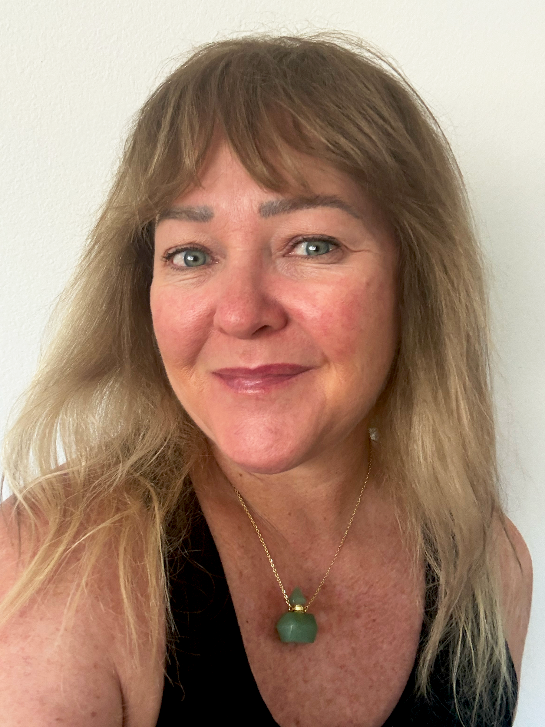 Julia Gomelsky standing in front of white wall wearing a black tank top and a green necklace smiling at camera
