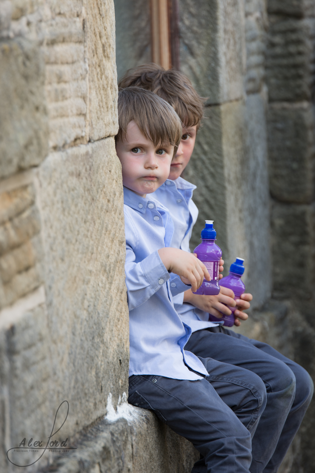 Two young boy wedding guests sit on a wall and have a fruit shot drink during the wedding reception welcome drinks