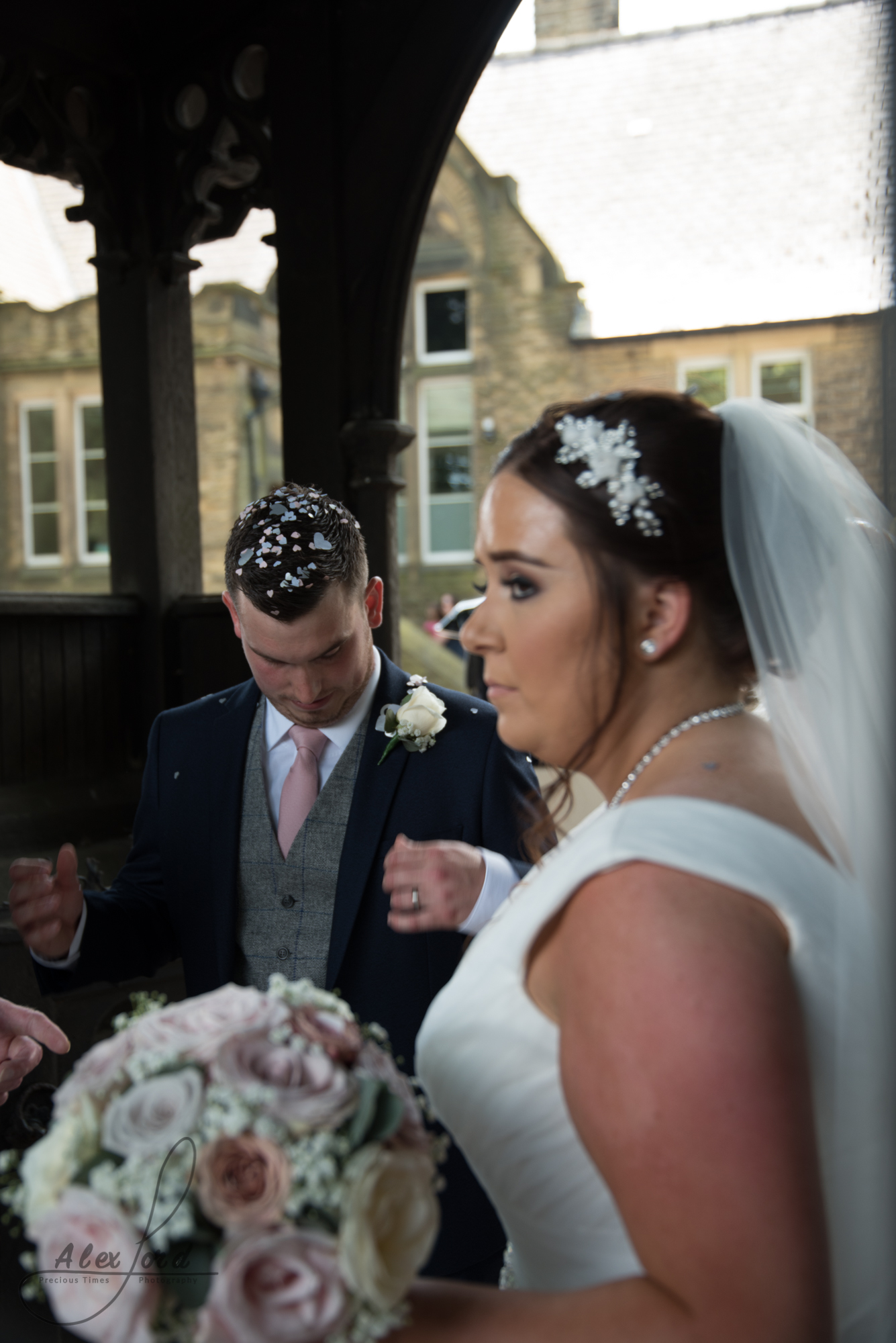 The bride and groom stand under and archway at the entrance to the church yard and wait for their guests to assemble for the confetti shot